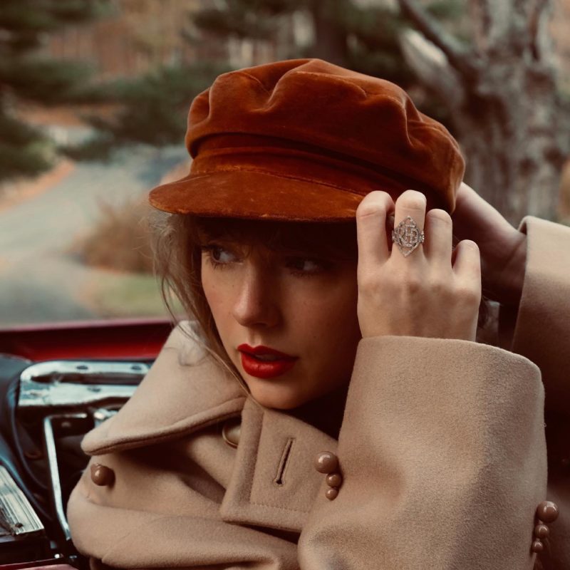 Capa do CD Red (Taylor's Versison). A foto mostra a cantora, uma mulher branca e loira, sentada dentro de um carro, usando chapéu vermelho, casaco bege e um anel com a palavra Red. Ela segura o chapéu com as mãos, ajustando-o na cabeça.