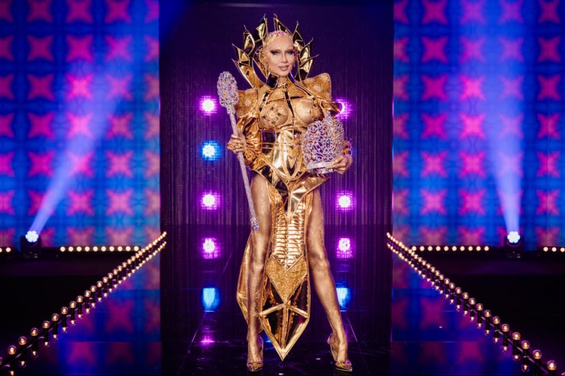 Cena de Drag Race Holland, mostra a drag Vanessa Van Cartier com a Coroa e o Cetro nos braços. Ela é uma mulher branca, que usa tinta dourada no couro da cabeça, além de um visual de gladiadora na mesma cor.