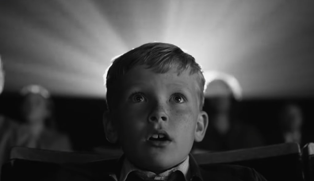 Cena do filme Belfast exibe, em preto e branco, um menino branco, de 9 anos, maravilhado ao assistir a um filme no cinema. Ele tem cabelo loiro, curto e está boquiaberto. Ao fundo, vemos a luz do projetor e os outros espectadores, desfocados. 