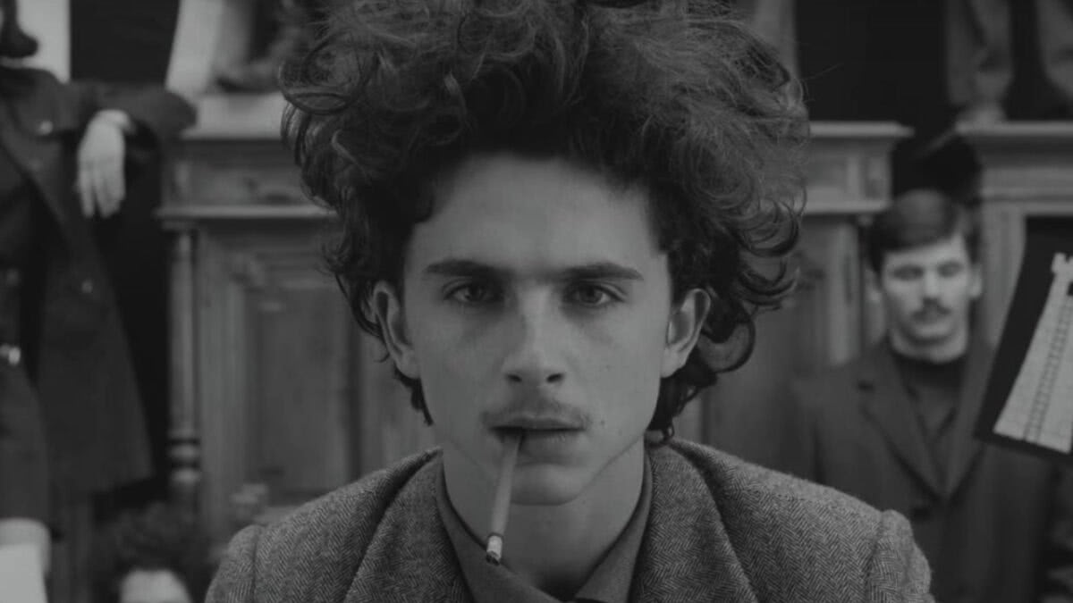 Cena de A Crônica Francesa. A imagem é um frame em preto e branco do rosto do ator Timothée Chalamet. Ele é um homem jovem e seu cabelo é liso. Há um cigarro em seus lábios.