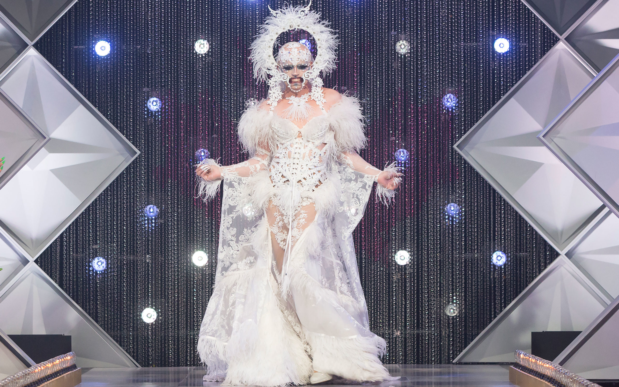 Cena da segunda temporada de Canada’s Drag Race, mostra a drag queen Icesis Couture desfilando pela passarela. Ela é branca, careca e usa roupas brancas, com uma espécie de aparelho bucal que abre sua boca mostrando a gengiva. O visual ainda tem uma auréola acima da cabeça.