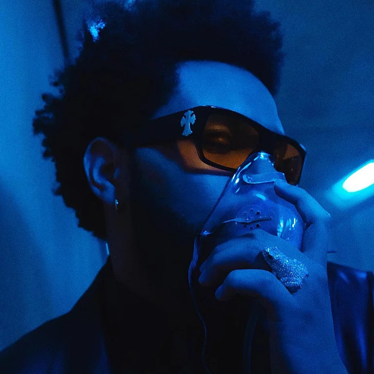  Imagem promocional de The Weeknd para o videoclipe de Take My Breath. Em tons azuis predominantes, a lente laranja do óculos de armação preta com uma cruz desenhada é a única peça que destoa dessa cor. O artista segura uma máscara de oxigênio com uma de suas mãos, na qual há também um anel em formato de sapo.