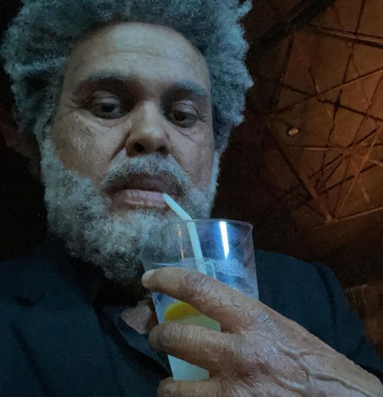 Selfie de The Weeknd caracterizado como um idoso de cabelo e barba grisalhos. O canadense de pele negra e olhos escuros, veste um terno preto enquanto ingere uma bebida em um copo transparente com canudo.