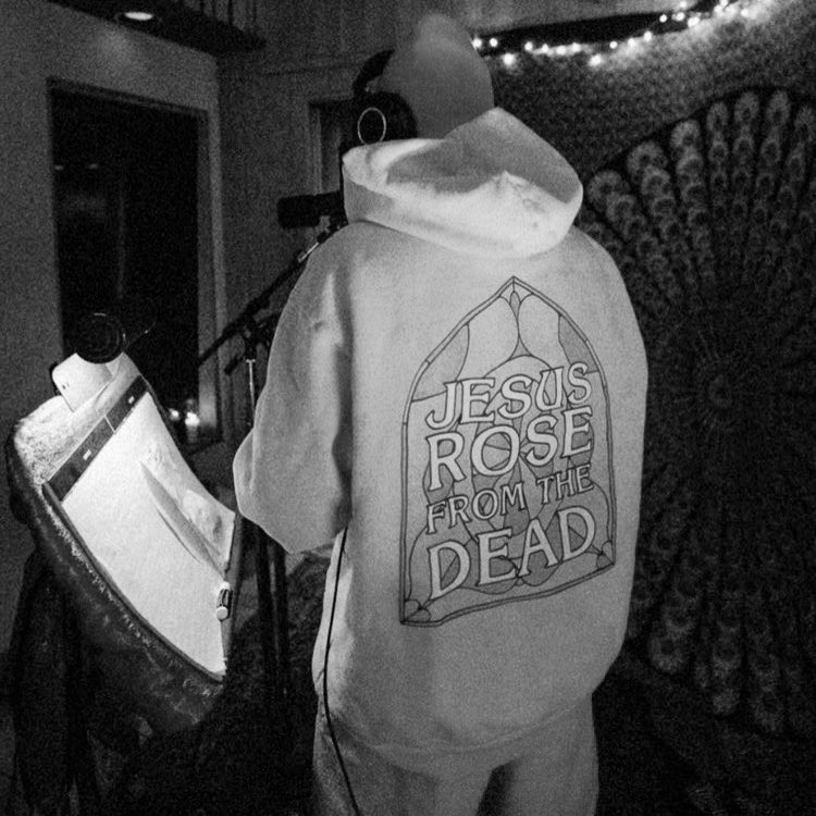 A foto está em preto e branco. Nela, o cantor Justin Bieber aparece de costas em um estúdio de música, usando um fone preto e vestindo um moletom escrito “Jesus rose from the dead”.