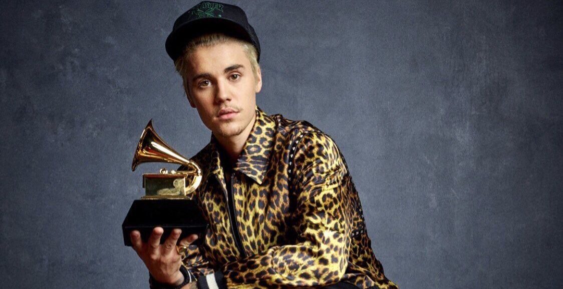Justin Bieber está posando para uma foto, segurando sua estatueta dourada do Grammy. Ele usa um boné preto e uma jaqueta com estampa de onça. 