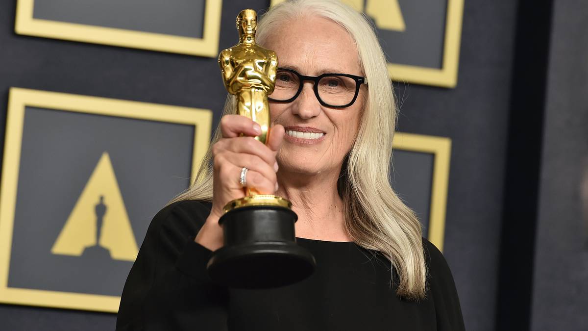 Foto da diretora Jane Campion, uma mulher idosa, de cabelos compridos grisalhos e óculos de grau com armação preta, segurando seu Oscar. Ela usa um vestido preto de manga longa.