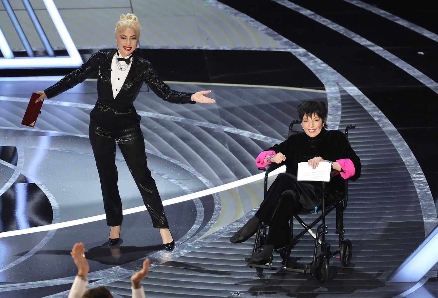 Foto das atrizes Lady Gaga e Liza Minnelli no palco do Oscar 2022. Gaga é uma mulher branca, usa um terno e tem os cabelos loiros presos num coque. Liza é uma mulher branca, idosa e está em uma cadeira de rodas preta, segurando um envelope branco.