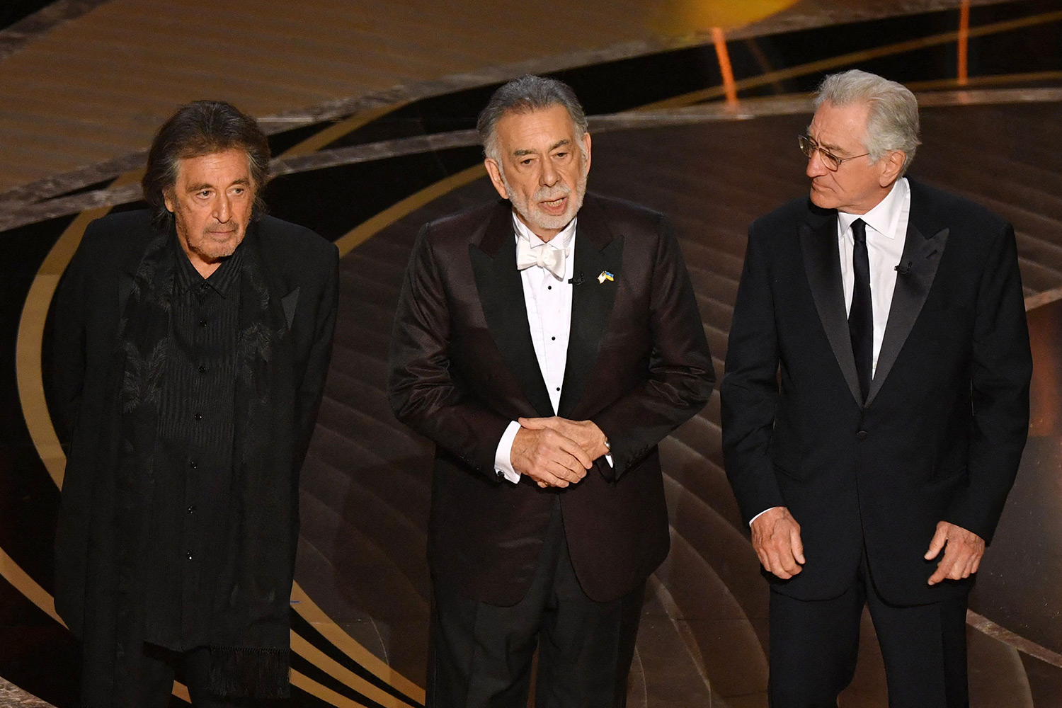 Foto do elenco de O Poderoso Chefão no palco do Oscar 2022. Na foto, vemos 3 homens idosos, todos de terno preto: Al Pacino, Francis Ford Coppola e Robert De Niro.