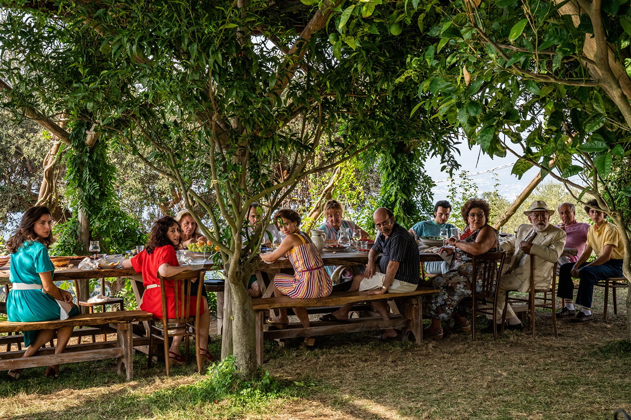 Cena do filme A Mão de Deus. A cena mostra uma família reunida ao redor de uma mesa grande, ao ar livre, rodeados por árvores em um belo jardim. Sentados em bancos, todos olham para algo fora da imagem, surpresos. 