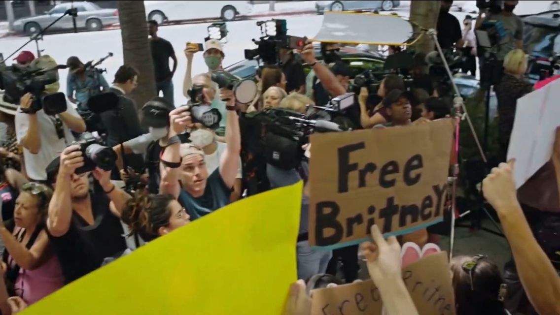 Cena do documentário Controlling Britney Spears: Em Busca de Liberdade. Na imagem, fotógrafos registram a manifestação de fãs da cantora do lado de fora do tribunal de justiça. No meio de vários cartazes, um se destaca: o pedaço de papelão escrito Free Britney (Liberte a Britney) em preto. Ao fundo, uma rua com carros de tons cinza.