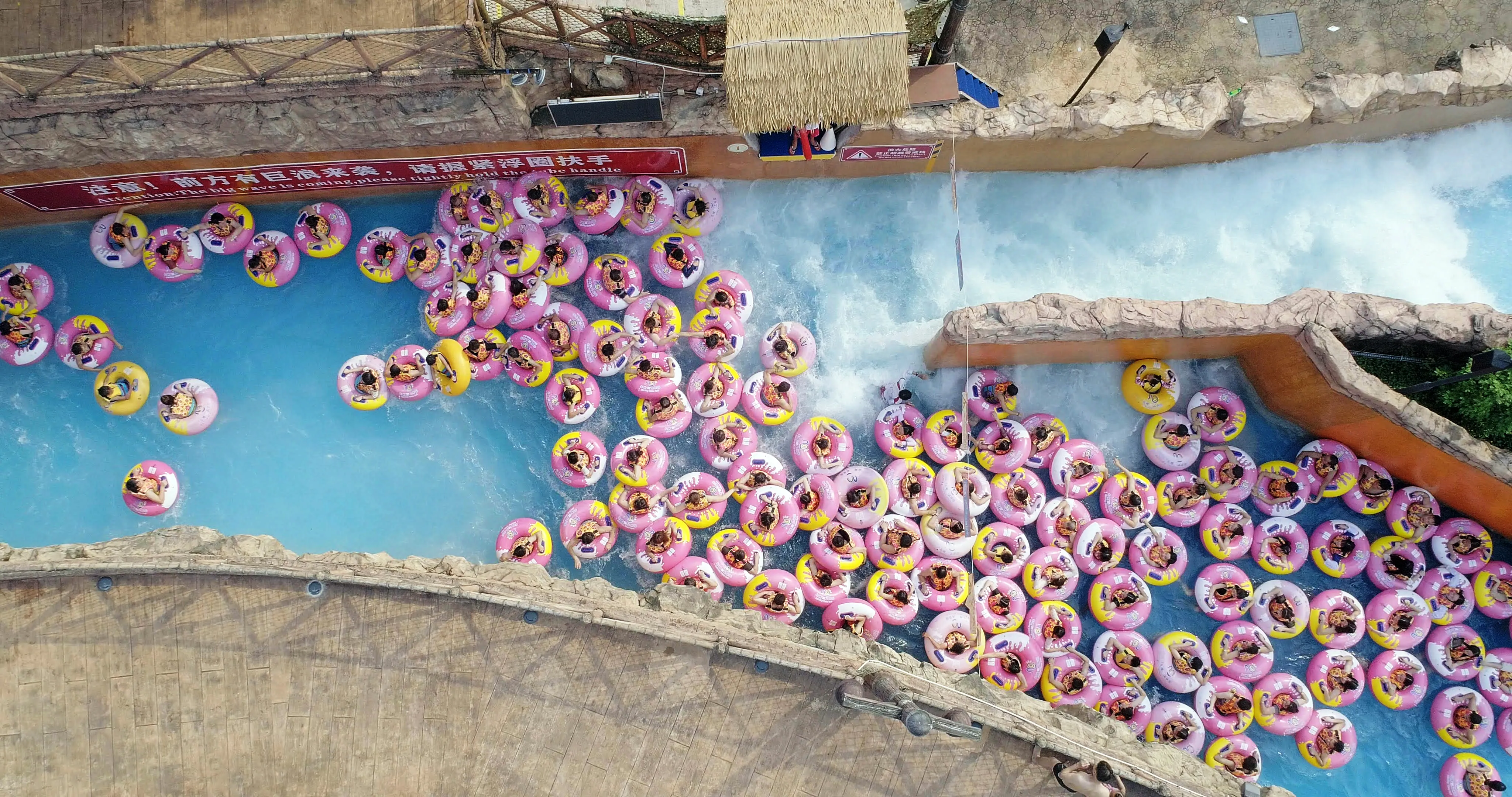 Cena do documentário Ascension. Imagem retangular e colorida. Nela, vemos de cima, um grupo enorme de pessoas, vestindo bóias rosas e amarelas, sendo levados pela correnteza de uma grande piscina de um parque aquático. O cenário é dia.