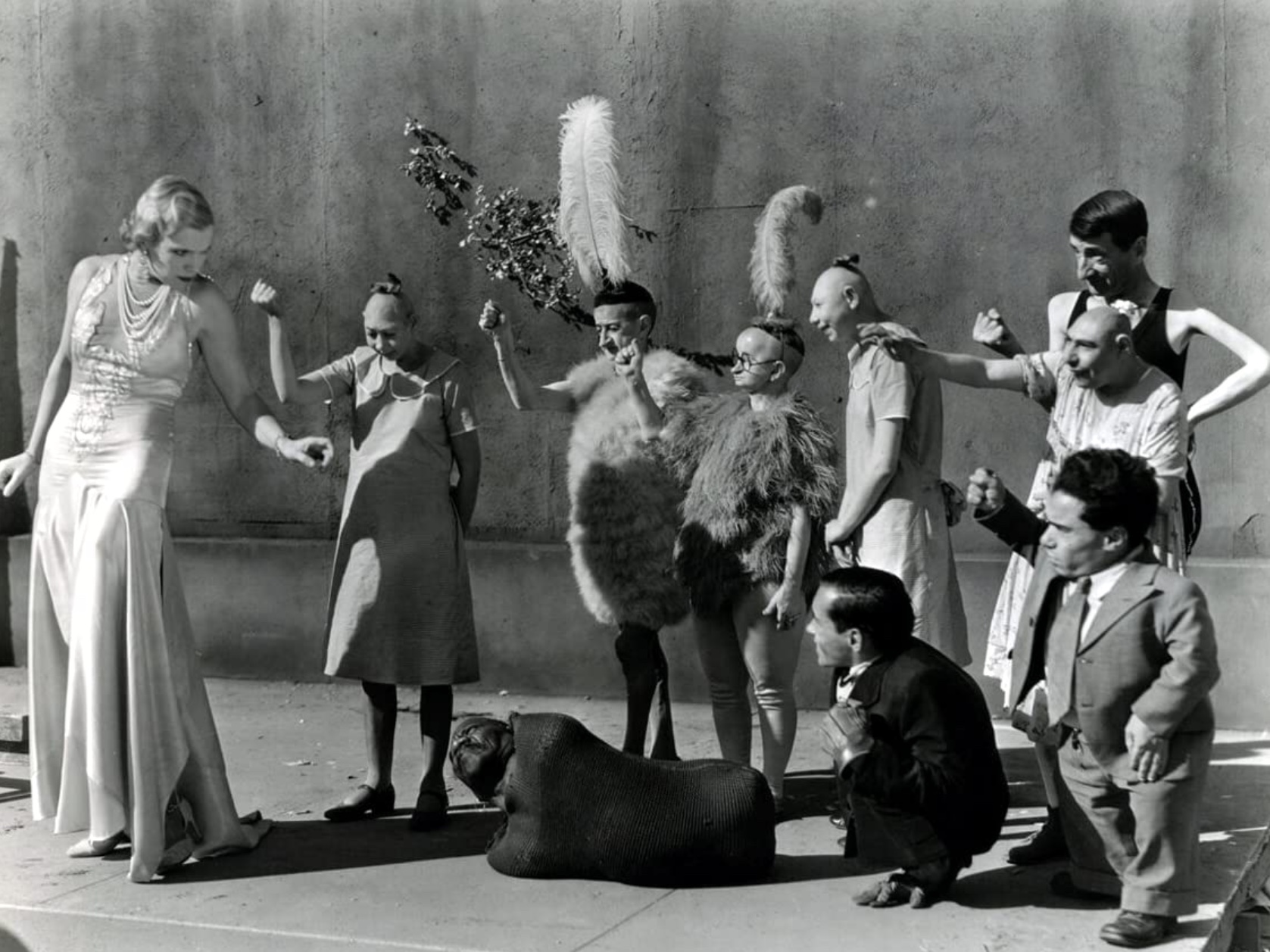 Cena em branco e preto do filme freaks apresenta um grupo de dez artistas de circo. Com exceção da mulher branca e loira, que usa um longo vestido branco e apresenta um olhar sério, todos os outros apresentam expressões de descontração.
