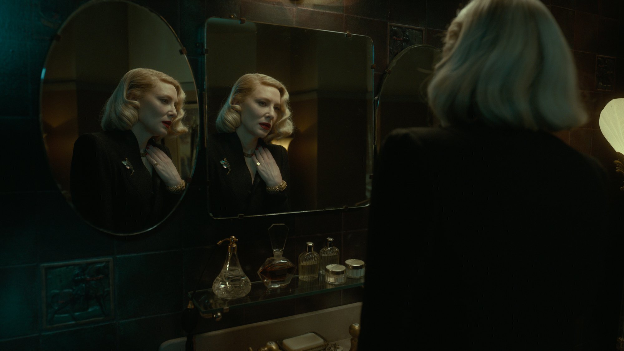 A imagem retangular é uma cena de O Beco do Pesadelo. À direita da foto vemos Cate Blanchett de costas da cintura para cima. Ela é uma mulher branca, mais velha, magra, de cabelos médios e encaracolados e bem loiros, que usa um batom vermelho vibrante. À sua esquerda vemos dois espelhos mostrando feições preocupadas olhando de volta para a atriz. O cenário é um banheiro escuro com pouca iluminação do lado direito da foto.