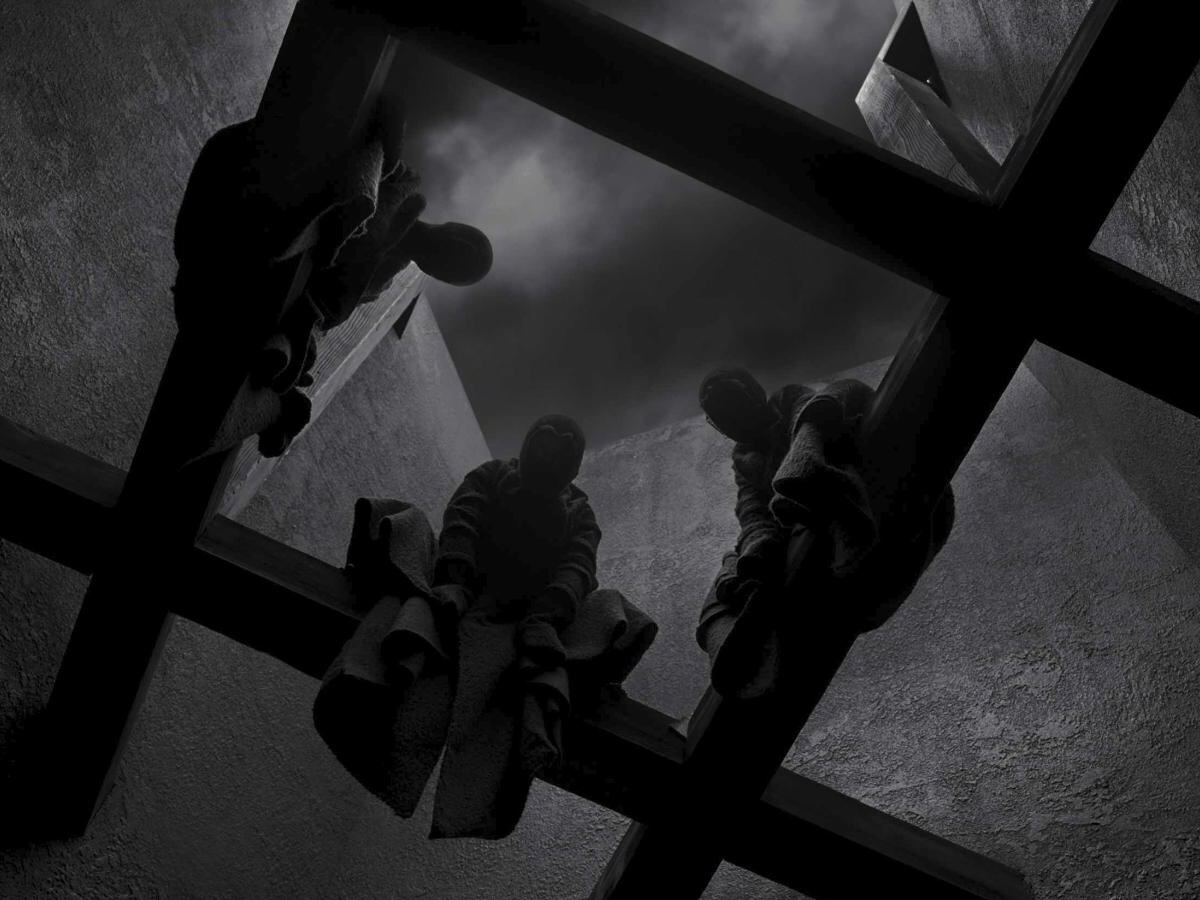 Cena em branco e preto do filme a tragédia de macbeth exibe três bruxas encapusadas com um longo manto negro sentadas em vigas. A imagem está enquadrada de baixo para cima e recebe pouca iluminação da noite.