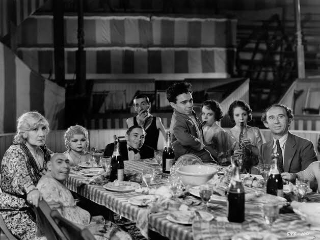Cena em branco e preto do filme freaks mostra um grupo de nove artistas de circo reunidos em volta de uma mesa bem comprida. Alguns dos personagens demonstram expressões de alegria e outros de tristeza. A mesa está coberta por uma toalha de piquenique e muita comida. A cerimônia se localiza em uma das tendas do circo.