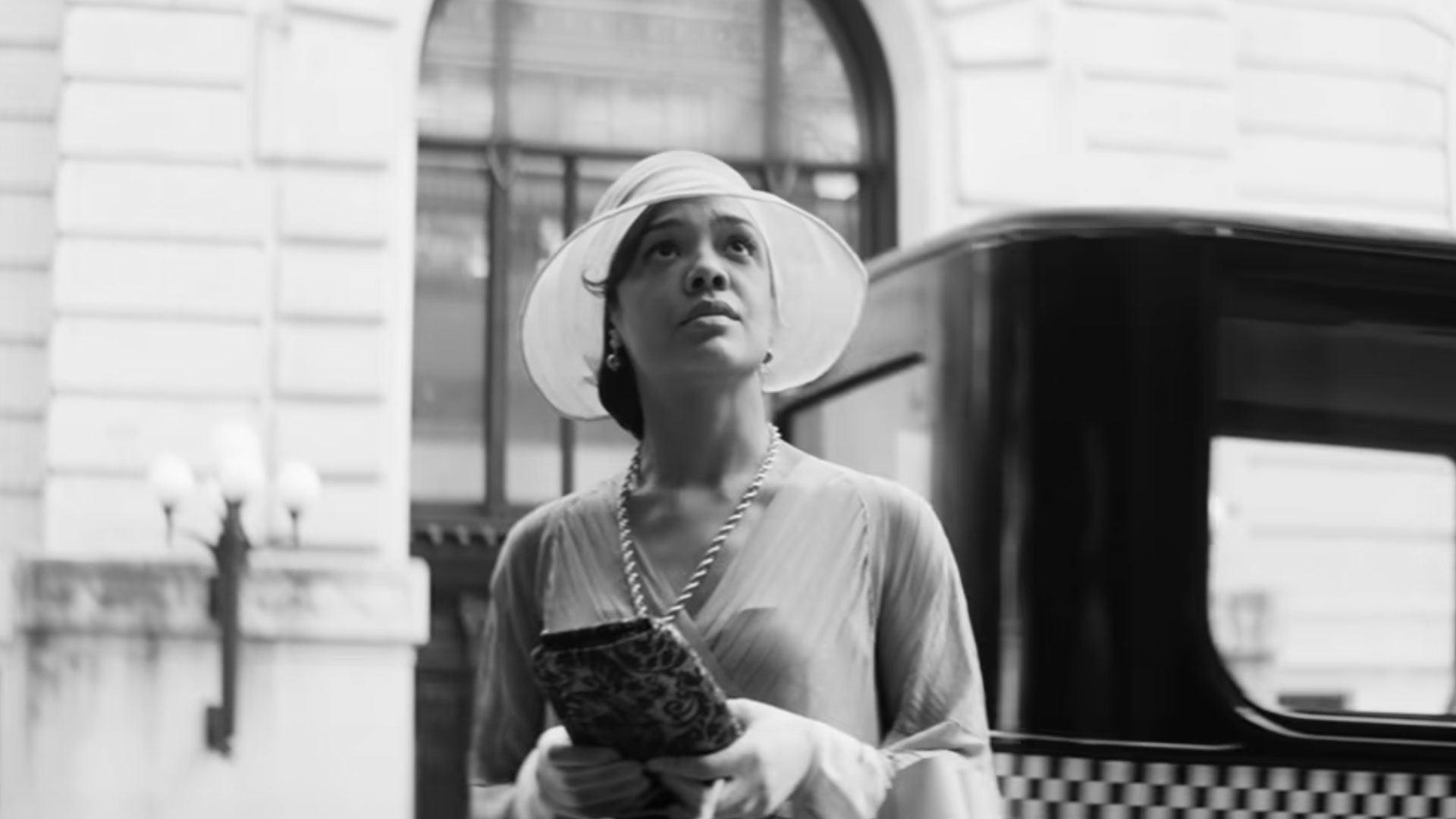 Cena do filme Identidade. Na imagem aparece Irene parada em frente a um ônibus, a  fotografia é em preto e branco. A personagem usa um chapéu e uma blusa de manga três quartos em tons claros, usa uma bolsa de mão e um colar de contas e segura sacolas de compras. 