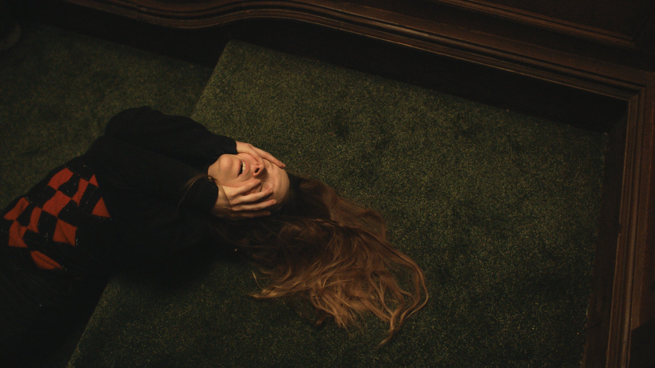 Cena do filme Saint Maud. A cena mostra a personagem Maud, uma mulher branca e de cabelos claros compridos, deitada no chão, com as mãos no rosto e o cabelo estirado atrás dela, como uma cascata. 