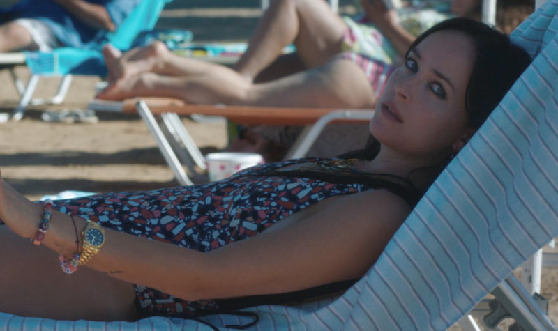 Cena do filme A Filha Perdida. Nela, está a atriz Dakota Johnson, que interpreta Nina, deitada em uma espreguiçadeira na praia, olhando para o lado. Nina é uma mulher branca, de cabelos escuros e compridos; ela veste um maiô colorido.