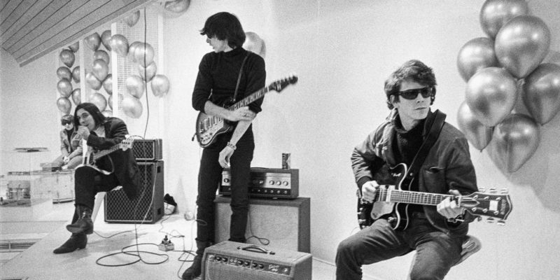Cena do filme The Velvet Underground. A cena mostra três músicos em um salão, rodeados de caixas de som. Todos são brancos, têm cabelo preto e usam roupas pretas com calça jeans. Estão com guitarras nos braços. Nas paredes, vemos vários balões. 