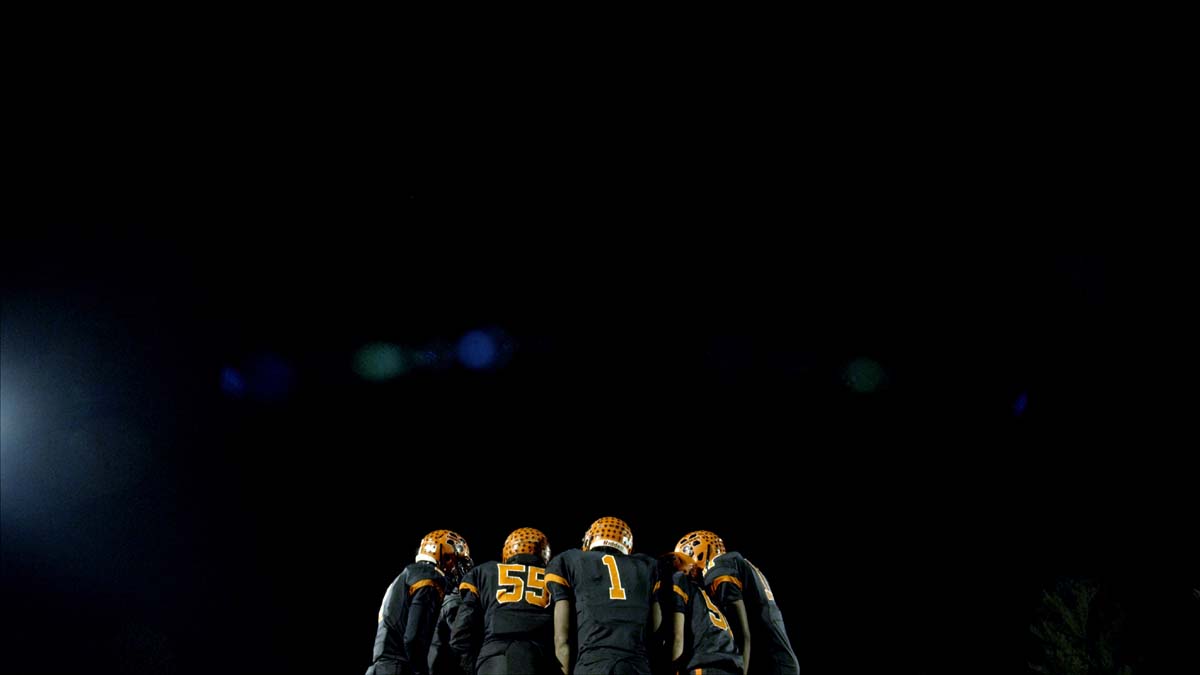 Cena do curta Audible mostra um time de futebol americano se reunindo antes do início do jogo, em um círculo fechado. Eles vestem roupas azuis e laranjas e o céu está preto atrás deles.