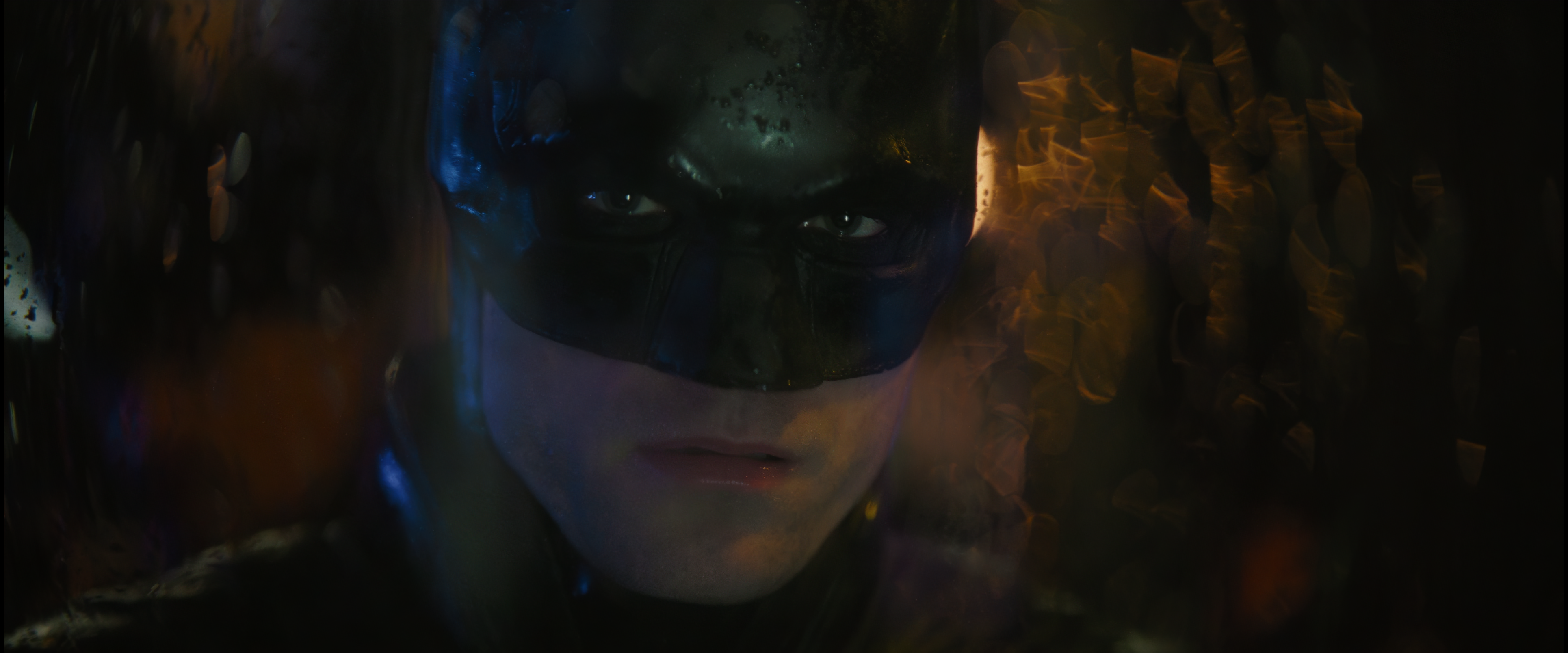 Cena do filme Batman. O Batman (Robert Pattinson) olha através de um vidro molhado, para frente. A metade superior de seu rosto é coberta por um capacete preto que mostra apenas os olhos. Ele usa uma capa preta com um colarinho alto. Ele é um homem caucasiano, de olhos azuis. Uma luz amarela vem detrás dele, iluminando as gotas no vidro, fora de foco. Está de noite e tons escuros e amarelos aparecem atrás dele, também fora de foco.