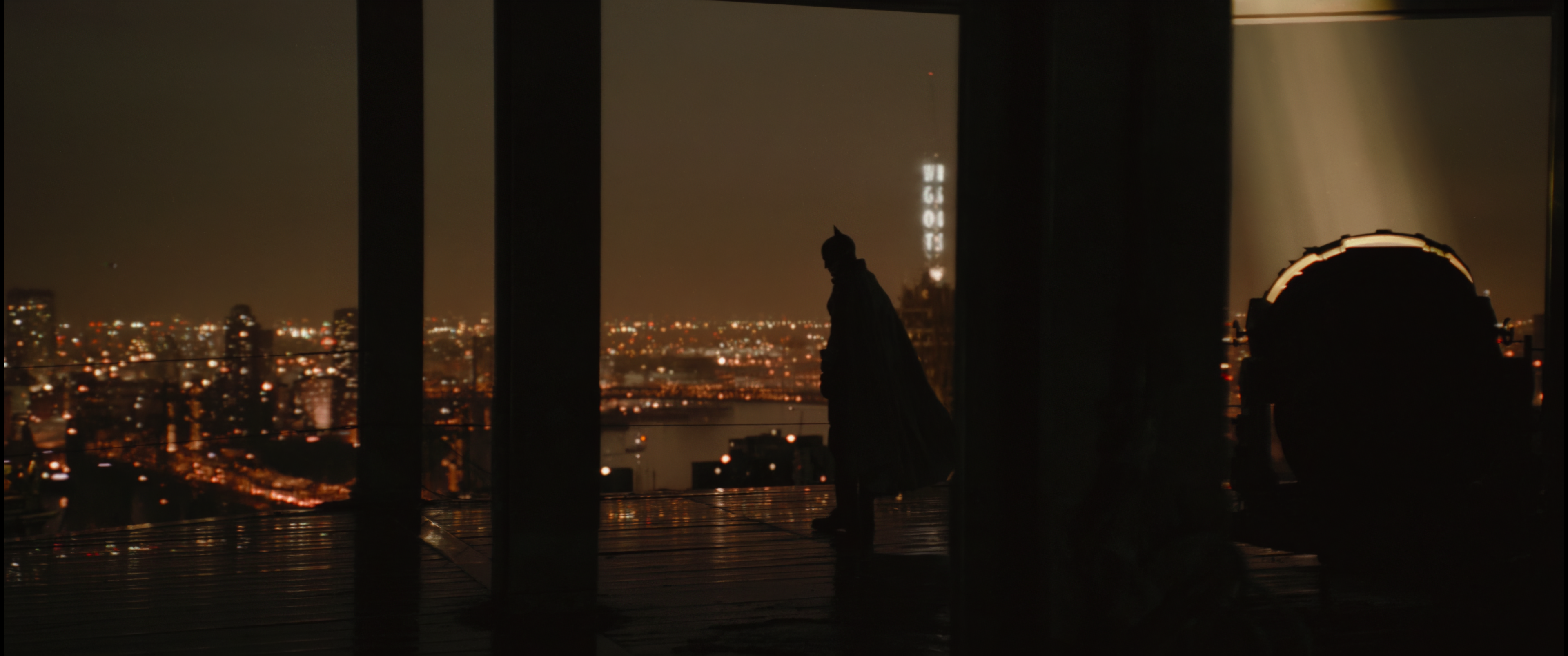 Cena do filme Batman. O Batman (Robert Pattinson) está no topo de um edifício em construção, virado para a esquerda, observando a cidade de Gotham à noite. Sua silhueta deixa evidente orelhas pontudas em seu capacete e uma capa esvoaçante para a direita. Atrás dele, à direita da tela e separado por vigas expostas, um holofote aceso aponta para o céu. Outras duas vigas menores, à esquerda, cortam a tela. O chão do edifício parece reflexivo e úmido. A cidade de Gotham é cortada por um rio e, ao longe, uma torre de rádio informa sua estação com um letreiro vertical luminoso.