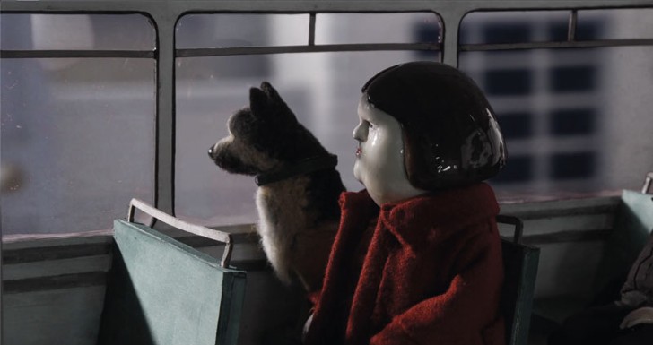 Cena do curta-metragem Bestia. Ao centro da imagem, uma animação, vemos a personagem Bestia sentada em um banco dentro de ônibus, ao lado de seu cachorro. Ambos olham pela janela do ônibus. O cachorro é um pastor alemão, usando uma coleira preta. A personagem é uma mulher, com a aparência de uma boneca de porcelana, com cabelos castanhos lisos na altura do queixo, vestindo um casaco vermelho.