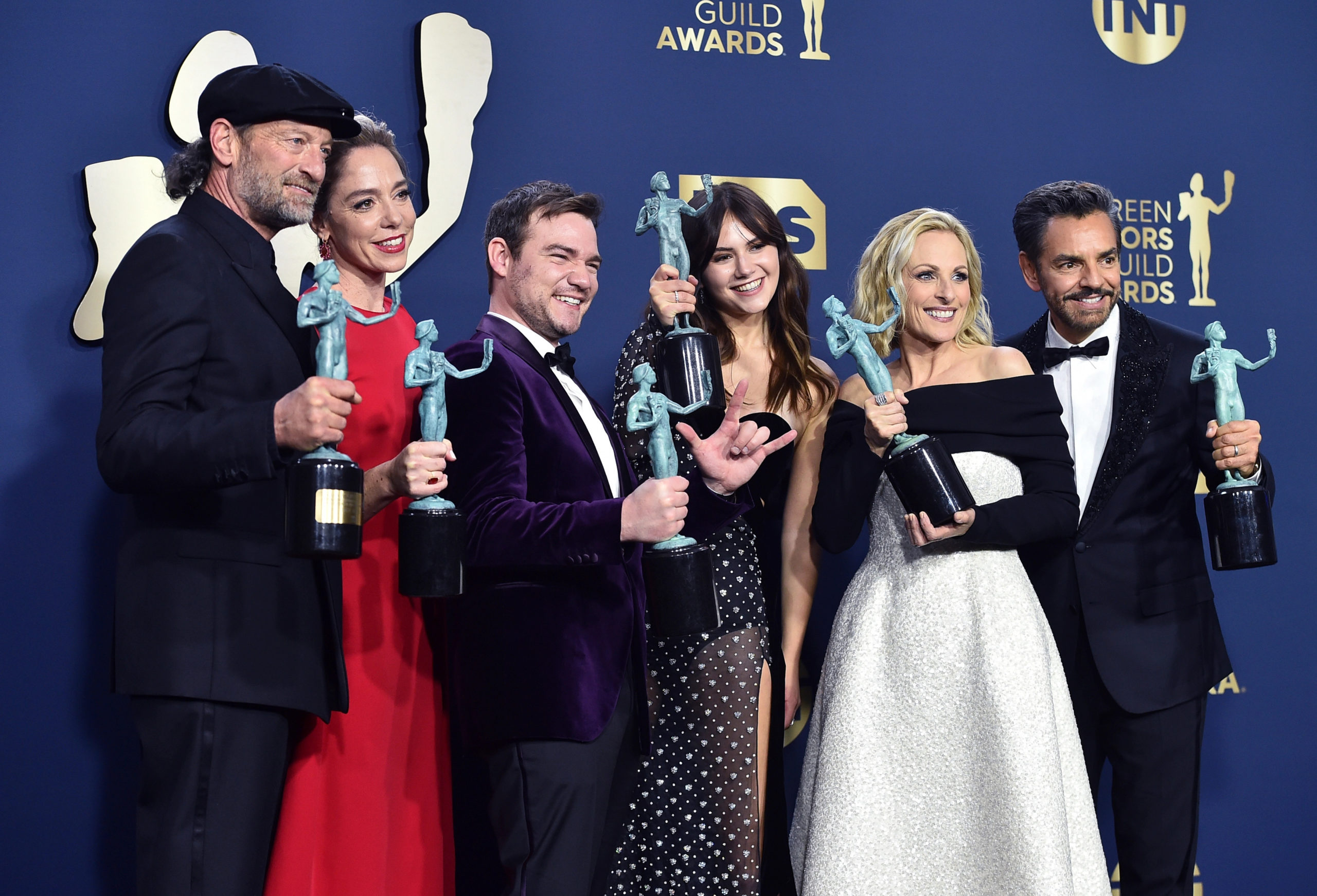 Foto do elenco do filme CODA no tapete vermelho do SAG Awards, com seus prêmios em mãos. Vemos 6 pessoas posando para uma fotografia, todos são pessoas brancas, e usam vestidos e ternos formais, segurando seus troféus e sorrindo.