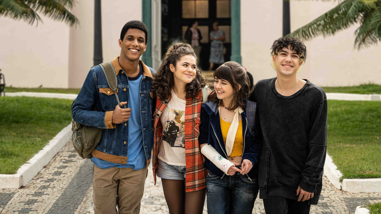 Cena da série De Volta aos 15. A imagem mostra um grupo de 4 amigos adolescentes saindo da escola, abraçados. É de dia e todos estão sorrindo. 