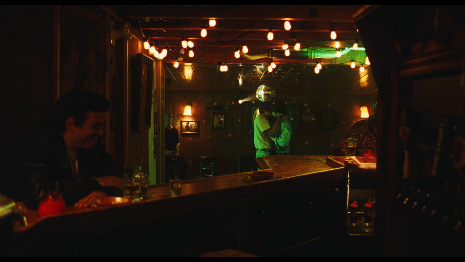 Cena da segunda temporada da série Euphoria. Na cena vemos um bar à noite e, no fundo da imagem, dois jovens adolescentes brancos se beijando. Eles se abraçam e se beijam e uma luz verde os ilumina. Na frente da imagem e desfocado, vemos um homem sentado no balcão do bar, sorrindo. 