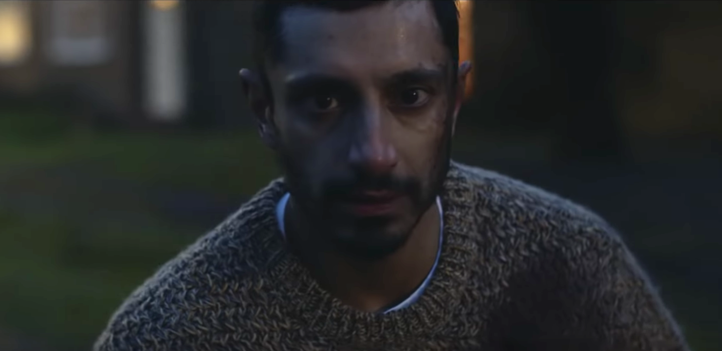 Cena do curta The Long Goodbye, mostra o ator Riz Ahmed, homem adulto de pele marrom, olhando para a câmera. Está anoitecendo e ele veste um suéter bege.