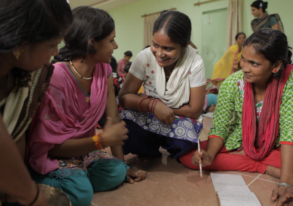 Cena do documentário Escrevendo com Fogo. A cena mostra 4 mulheres indianas sentadas no chão, escrevendo num papel e sorrindo umas para as outras.