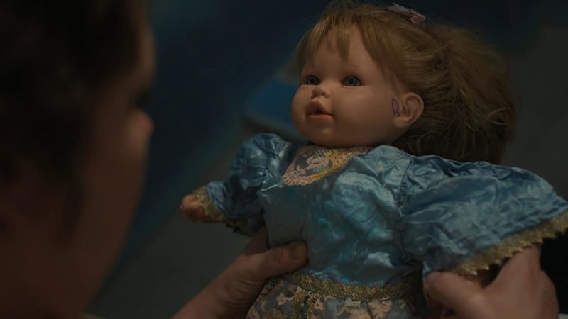 Cena do filme A Filha Perdida. Nela, vemos uma boneca, que está sendo segurada pelas mãos de Leda. A boneca é branca, com cabelos castanhos claros e veste um vestido azul claro.