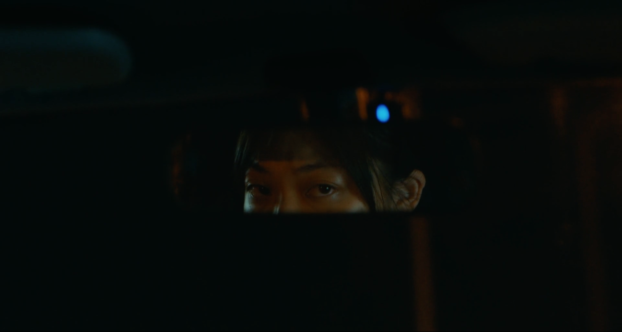 Cena do filme Drive My Car. Nela está o reflexo do olhar de Misaki pelo retrovisor. Misaki é uma jovem nipônica com cabelos pretos. O fundo do retrovisor é o céu da noite, completamente preto.