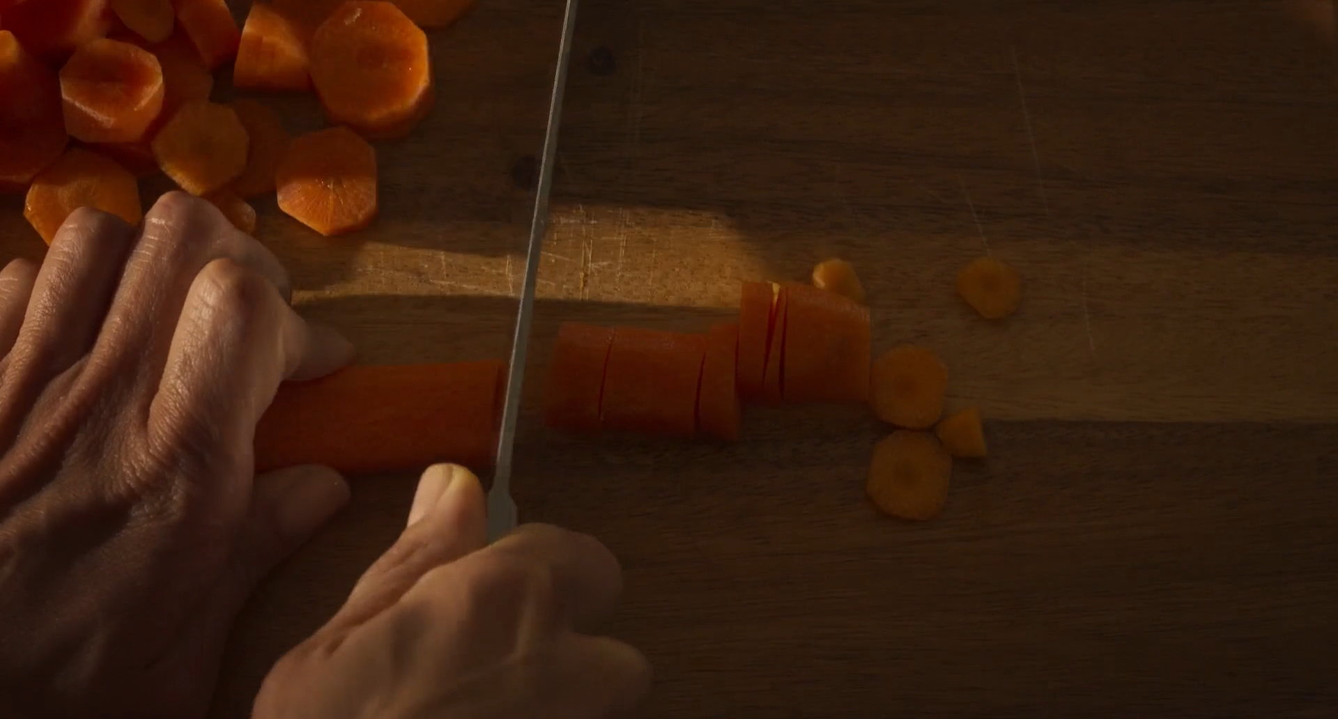 Cena do filme Mães Paralelas. Nela aparece um par de mãos cortando uma cenoura laranja com uma faca de inox prateada. As mãos estão ampliadas. O fundo é uma tábua de madeira com marcas de usos.