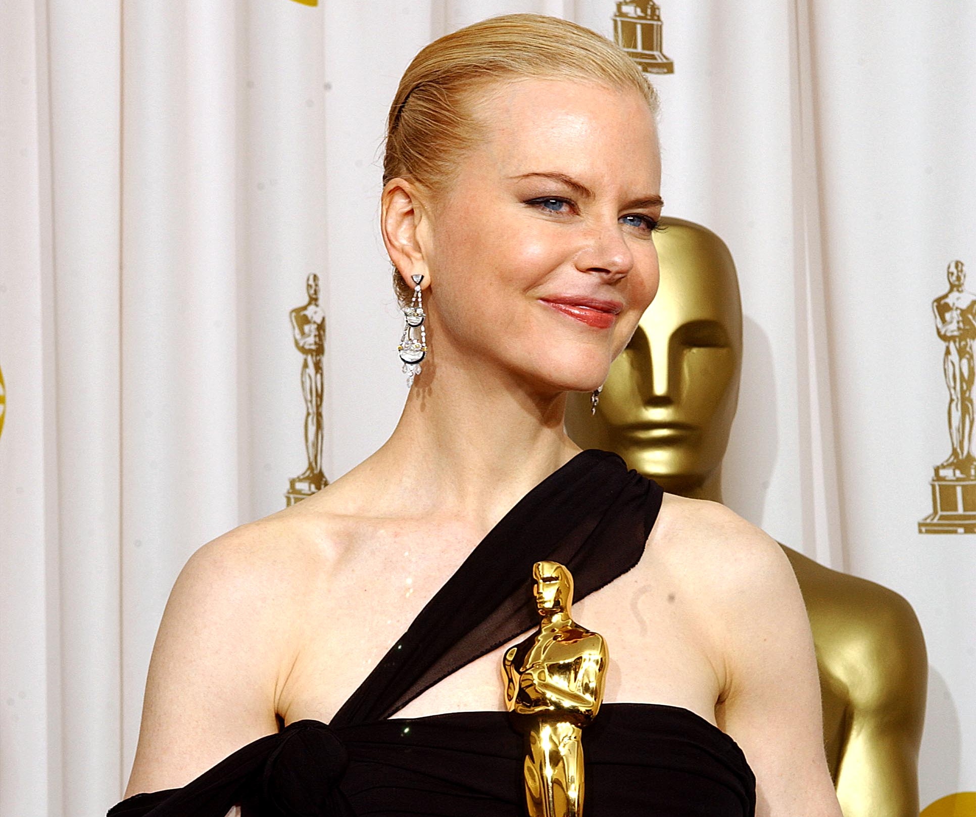 Foto da atriz Nicole Kidman segurando uma estatueta do Oscar. Nicole é uma mulher branca de olhos azuis, ela tem cabelos loiros, que estão presos em um coque baixo; ela veste um vestido preto com uma alça atravessada no colo do seu peito e usa um par de brincos com pedras. Ao fundo, é possível ver uma parte da estatueta do Oscar em tamanho grande. 