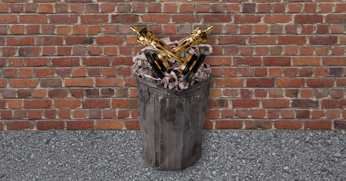 Arte com uma lata de lixo preta parada em frente a uma parede de tijolos laranjas. Dentro da lata de lixo, vemos duas estatuetas douradas do Oscar, deitadas em formato de X.