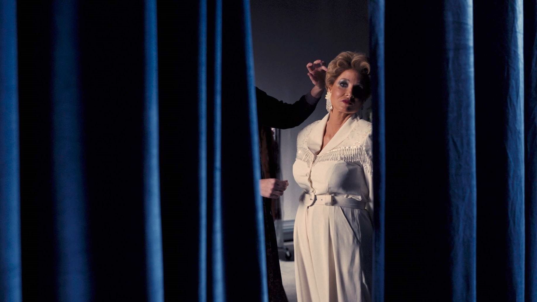Cena do filme Os Olhos de Tammy Faye, mostra a personagem principal se olhando por entre duas cortinas azuis. Ela é uma mulher branca, adulta e usa roupas brancas.