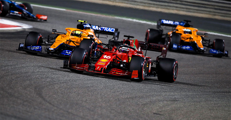 Foto de três carros de Fórmula 1. Na imagem, dois carros da equipe McLaren, nas cores mamão-papaia com detalhes em azul, perseguem um carro vermelho da equipe Ferrari. O ambiente é uma pista de corrida cinza.