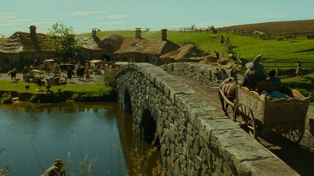 Cena do filme O Senhor dos Anéis: A Sociedade do Anel. Gandalf e Frodo chegam no Condado em uma carroça movida por um cavalo marrom. Ela começa a cruzar uma ponte de pedra, da direita para a esquerda, em direção a um amontoado de casas, à esquerda da tela, com o rio que passa embaixo da ponte refletindo-o. O vilarejo se encontra no meio de um campo verdejante e, ao longo do horizonte, podemos ver alguns cavalos pastando na parte direita da tela. O céu é azul e sem nuvens. Gandalf usa um robe cinzento e um chapéu pontudo também cinzento enquanto só o topo da cabeça de Frodo é visível, os cabelos pretos encaracolados.