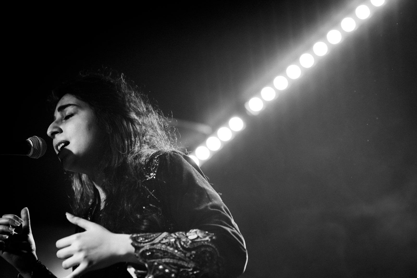 Foto em preto e branco da cantora Arooj Aftab em uma apresentação. Ela está no canto esquerdo da imagem, de perfil, cantando com um microfone, que está preso em pedestal, bem próximo a sua boca. Arooj é uma mulher de traços árabes e cabelos lisos compridos. Ela usa uma roupa de mangas compridas, com detalhes nas mangas. 