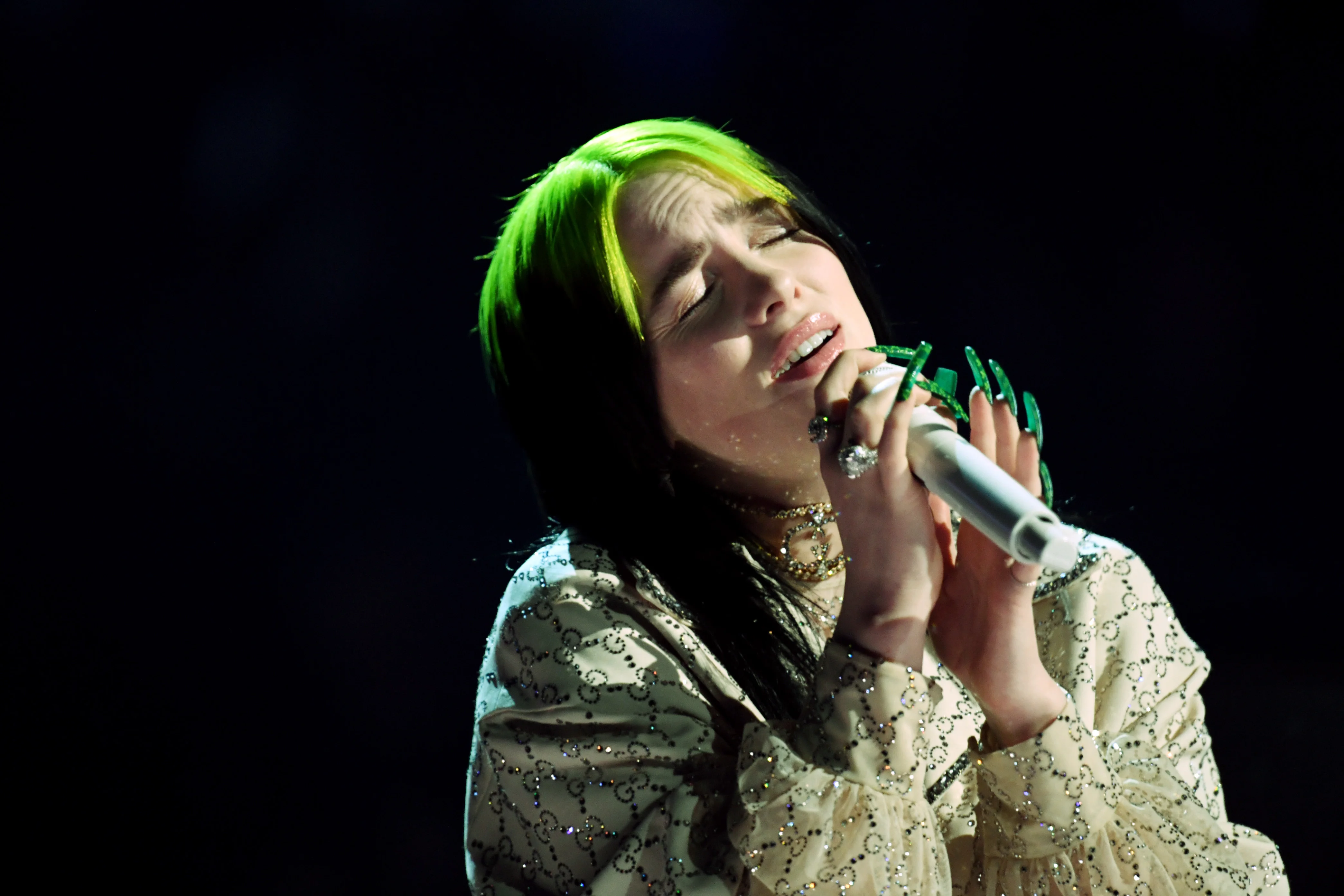 Foto de Billie Eilish cantando. Nela, a cantora está segurando um microfone na mão esquerda, próximo a sua boca. Billie é uma mulher branca, de cabelos pretos, mas a raiz pintada na cor verde neon. Ela veste um a blusa bege de mangas compridas e tem unhas compridas com esmalte verde.