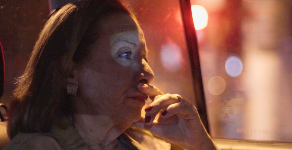Cena do filme Eneida, mostra a personagem titular pensativa, iluminada pelas luzes do trânsito à noite, olhando pela janela do carro. Ela é uma mulher branca, idosa, de cabelos claros e unhas vermelhas.
