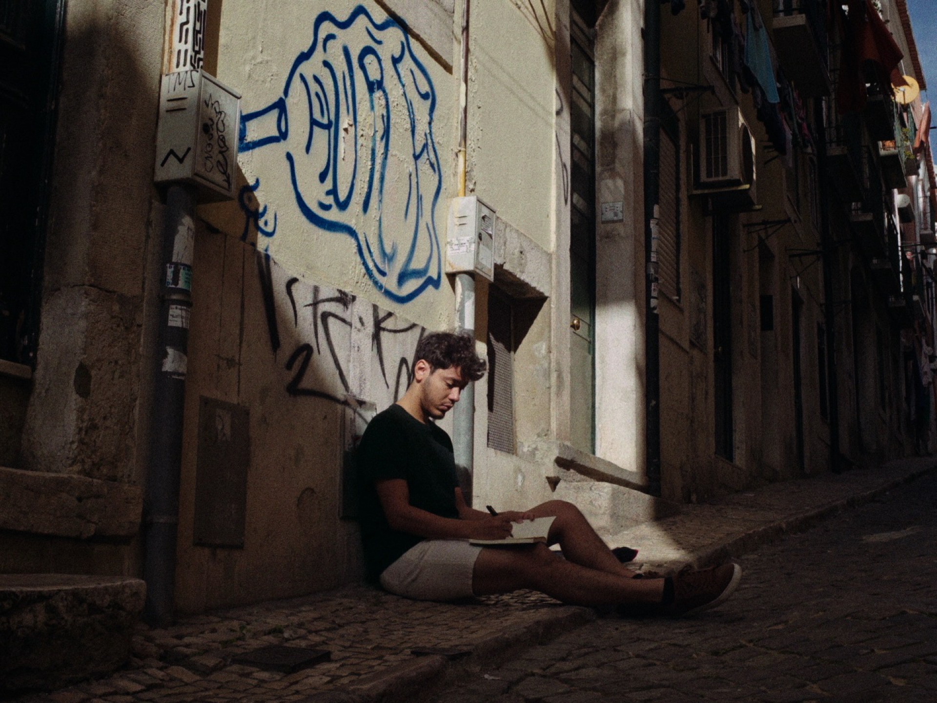Cena do curta Meio Ano-Luz, mostra um homem branco sentado à rua, com um caderno em mãos, desenhando. Atrás dele, a parede é cor de creme e tem uma pichação azul. 