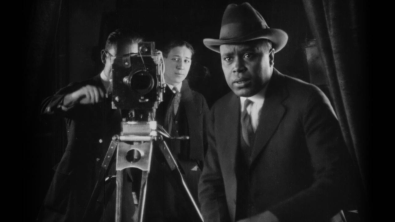 Cena em preto-e-branco do filme Oscar Micheaux: O Super-Herói do Cinema Negro, em que um homem negro, vestindo um terno e chapéu, olha para frente com atenção. Logo atrás dele, vê-se outros dois homens. Um deles manuseia uma câmera de filmagem antiga, e o outro, mais ao fundo, observa os dois.
