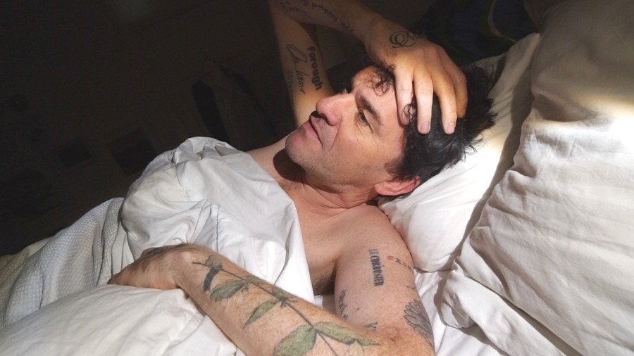 Cena do filme A História do Olhar, mostra o diretor Mark Cousins deitado na cama sem camisa. Ele é um homem branco, tem tatuagens nos braços e leva a mão direita à testa, olhando para o teto. 
