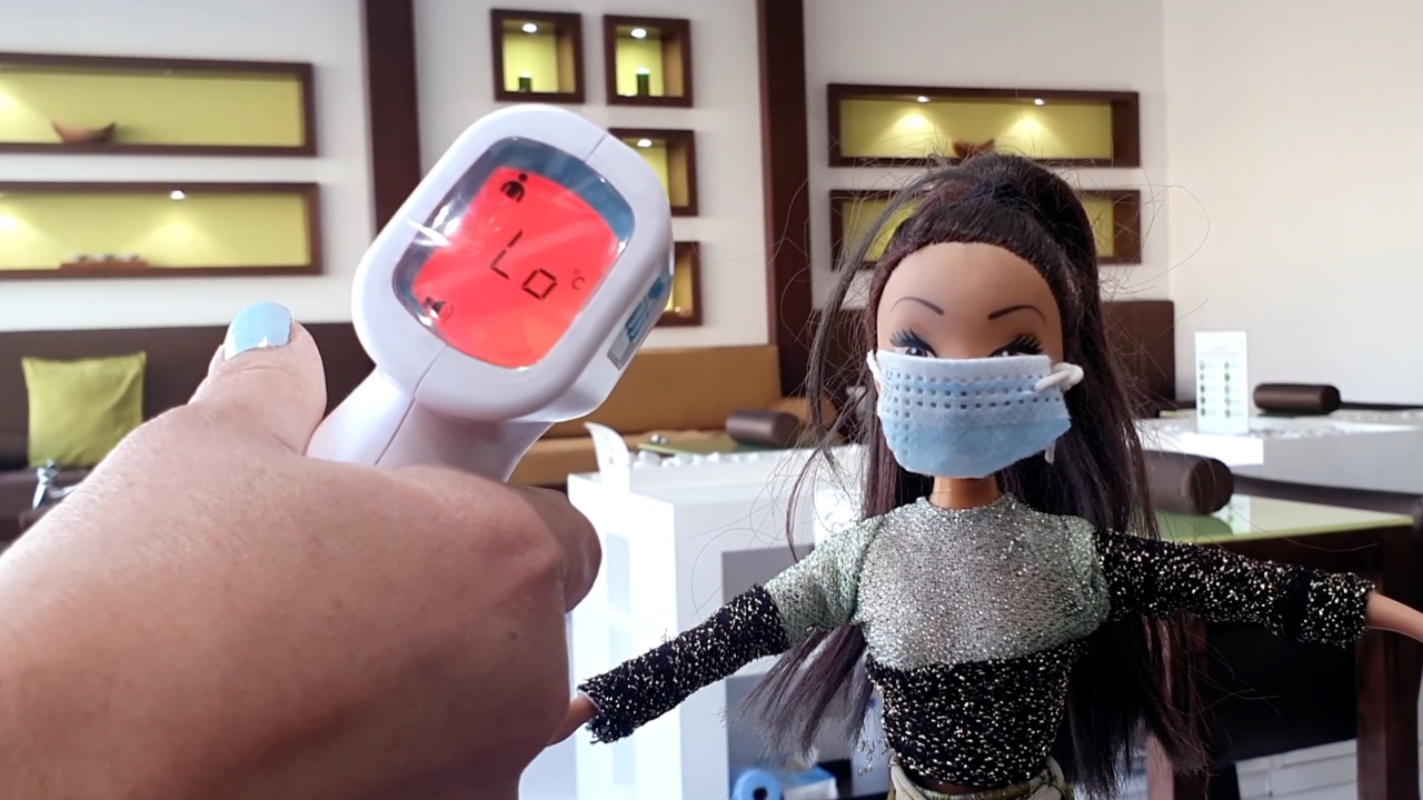 Cena do curta Voz Virtual, mostra uma boneca de máscara de pano tendo a temperatura medida na testa.