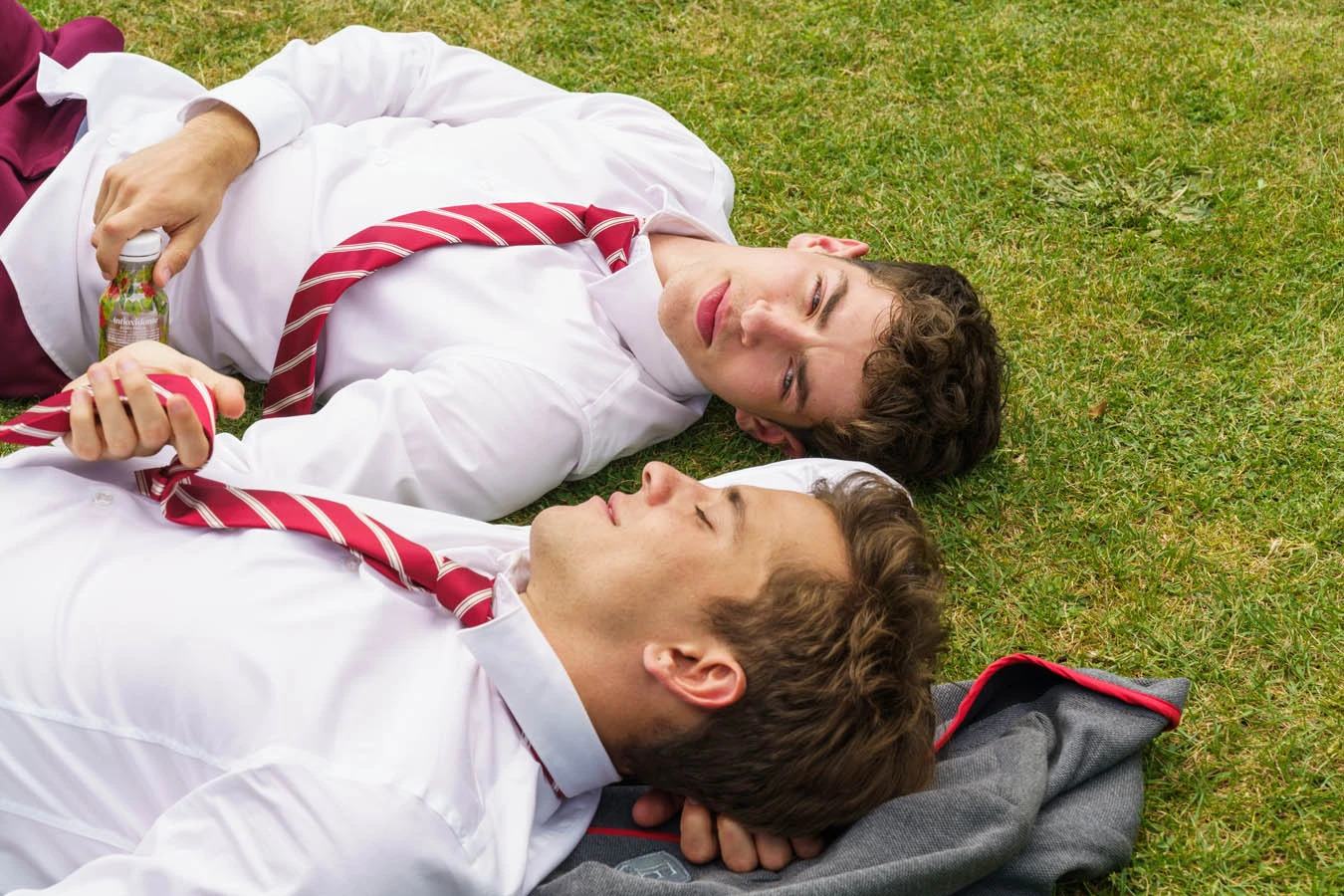 Cena da série Elite, mostra dois homens brancos deitados num gramado, sorrindo e se olhando. Eles usam camisas brancas, calças vermelhas e gravatas brancas e vermelhas. A grama é verde e está de dia.