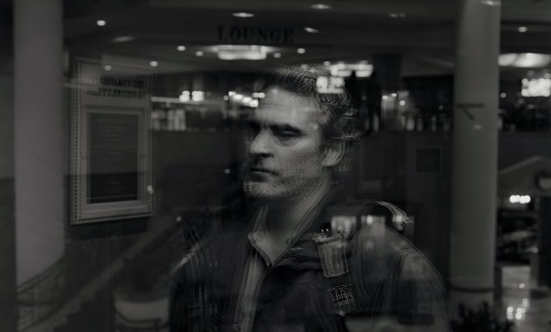 Cena do filme C’mon C’mon. É o reflexo de Johnny, interpretado por Joaquin Phoenix, em uma janela de vidro. Sua aparência é de um homem barbudo, já com cabelo branco e leves rugas no rosto. Está com um agasalho e mochila nas costas. Seu olhar é melancólico e contemplativo. 