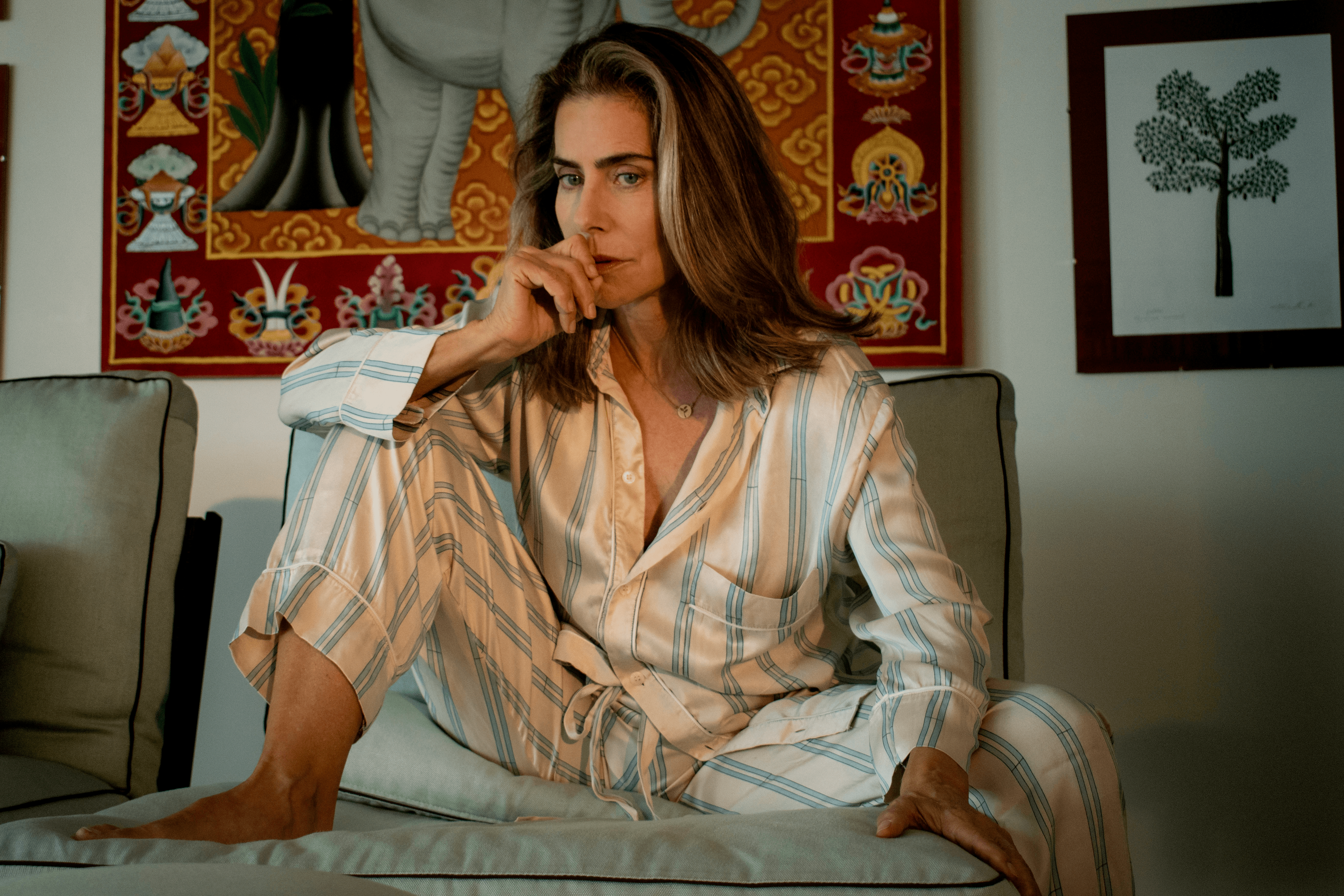 A imagem apresenta a atriz Maitê Proença, uma mulher branca e loira, que veste um pijama branco com listras azuis. Ela está sentada em um sofá cinza e podem-se ver um quadro indiano e um quadro de uma pequena árvore na parede do fundo